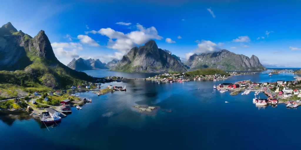 Panoramatický pohled na rybářskou vesnici v Lofotenských ostrovech, kde se loví ryby pro výrobu produktů Norsan Omega 3, obklopenou majestátními horami a klidnou modrou vodou.