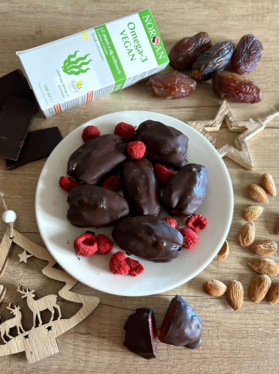 datle-v-cokolade-s-malinovym-kremem-norsan-omega-3-vegan Datle v čokoládě s malinovým krémem na talíři obklopené malinami, mandlí a produktem Norsan omega-3 Vegan.
